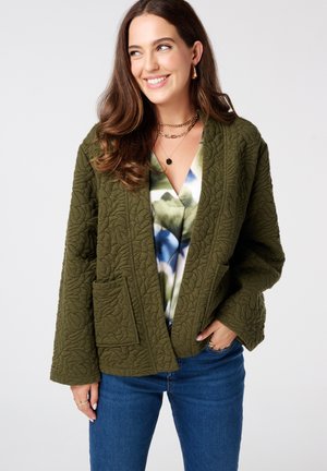 Veste en olive matelassée avec une coupe décontractée, présentant un décolleté en V profond, deux poches avant et un motif de feuilles texturé. Portée sur un haut à motifs floraux.