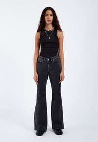 Vrouw met lang krullend haar, gekleed in een zwart mouwloos topje, hoog getailleerde donkere wijde jeans, zwarte schoenen en laagjes halskettingen, staand tegen een witte achtergrond.