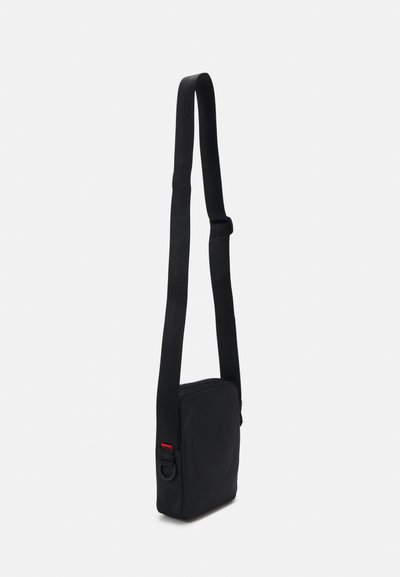 Sac bandoulière noir de forme carrée, fabriqué en matériau lisse. Il dispose d'une sangle réglable et d'un petit détail d'accent rouge.