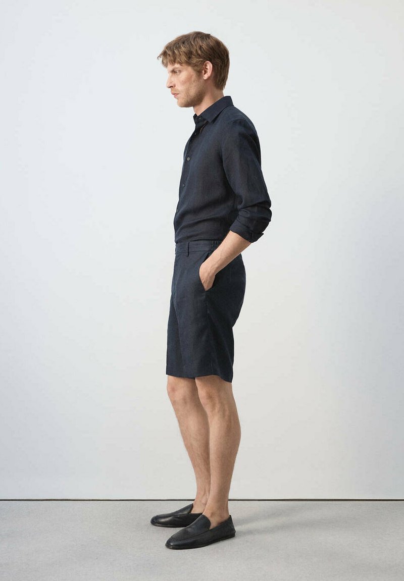Homme debout de profil, portant une chemise sombre à manches longues, un short assorti avec les mains dans les poches, et des chaussures noires sans lacets sur un fond uni.