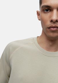 Beige sweatshirt featuring round neckline, subtle text detail on chest, and contrasting stitching along the shoulder seams. Smooth texture.