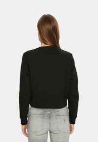 Felpa nera corta con scollatura a girocollo e polsini a coste, mostrata da dietro. Indossata con jeans attillati chiari.