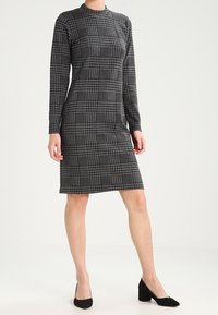 Robe moulante avec un motif pied-de-poule en noir et blanc, manches longues, col montant et un tissu lisse et extensible. Chaussures à talons noirs.