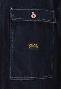 Seção de uma jaqueta de ganga azul escura com um bolso no peito com costura branca e um patch de logótipo preto que diz "Stan Ray" em dourado.