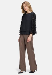 Blusa nera con maniche trasparenti a strati decorate con piume, abbinata a pantaloni larghi marroni e scarpe nere con punta affusolata.