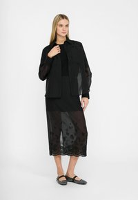 Giacca a camicia nera traslucida con chiusura a bottoni, indossata sopra un top nero e una gonna nera trasparente con ricami intricati e orlo scallop.
