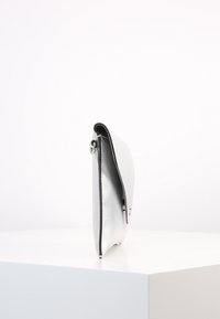 Srebrna metaliczna kopertówka o eleganckim, minimalistycznym wzornictwie, z gładką powierzchnią, zaokrąglonym kształtem i czarnym wykończeniem wzdłuż krawędzi.