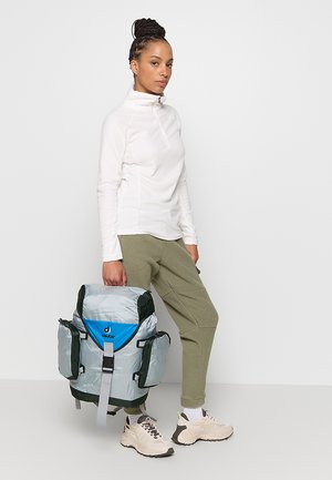 Lichtgrijze rugzak met donkergroene accenten, een gesp aan de voorkant en een blauw logo. Het model draagt een witte top en olijfgroene broek met beige schoenen.