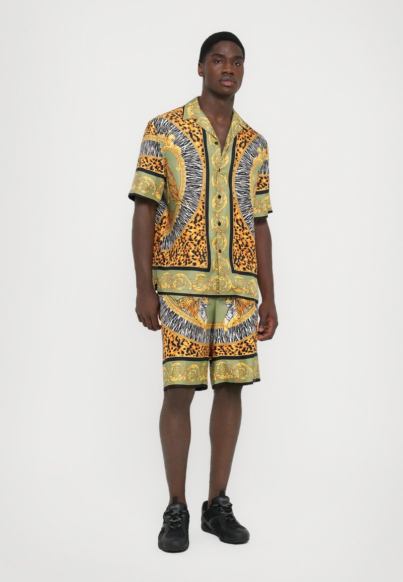 Korte mouwen overhemd en shorts set, met een groen, zwart en gouden dierenprint patroon, met een ontspannen pasvorm en een knoopsluiting aan de voorkant. Zwarte sneakers gedragen.