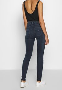 Dunkelblaue Skinny-Jeans mit hoher Taille, featuring einer glatten Oberfläche und subtilen Nähdetails. Kombiniert mit einem schwarzen Tanktop und weißen Sneakern.