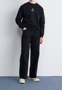 Black sweatshirt with a geometric emblem, paired with black straight-leg jeans and white sneakers. Fabric appears soft and matte.