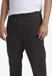 Pantaloni neri in tessuto elasticizzato con una texture liscia, dotati di coulisse e tasche laterali, mostrati dalla vita fino alle ginocchia.