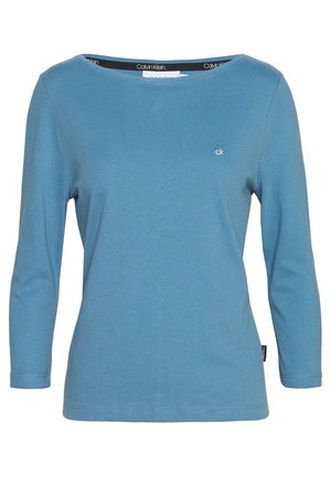 Niebieska, długorękawowa koszula Calvin Klein z okrągłym dekoltem i małym logo "ck" na piersi, jednolity wzór, widoczna metka marki wewnątrz kołnierza.