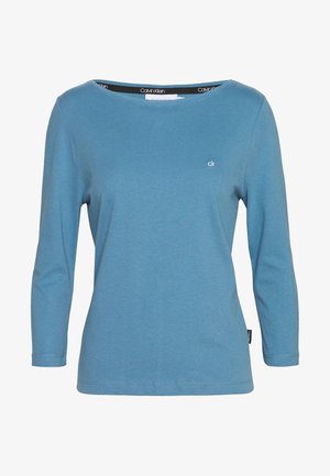 Modrá košile Calvin Klein s dlouhým rukávem, kulatým výstřihem a malým logem "ck" na hrudi, jednoduchý design, viditelná značka uvnitř límce.