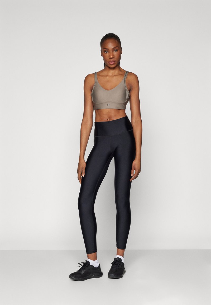 Sujetador deportivo de tela gris claro con tirantes ajustables, combinado con leggins negros de cintura alta. La modelo lleva zapatillas deportivas negras.