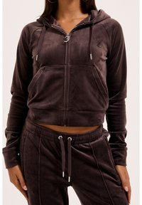 Femme portant un sweat à capuche zippé en velours brun foncé et un pantalon à cordon assorti sur fond uni.