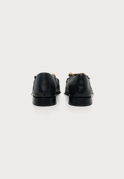 Paire de mocassins en cuir noir vus de dos avec une petite décoration en barre métallique dorée sur le dessus, sur fond blanc uni.