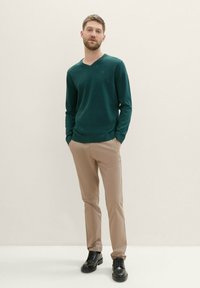 Pulover de culoare verde închis cu decolteu în V, pantaloni bej și pantofi negri cu șireturi. Textură moale, design ajustat și fundal neutru.