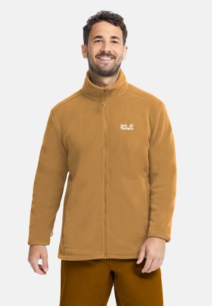 Uomo sorridente che indossa una giacca in pile marrone chiaro con zip e logo Jack Wolfskin sul petto e pantaloni marroni abbinati.