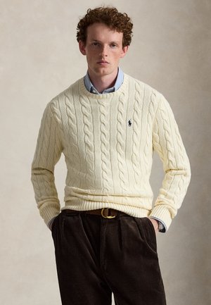 Un hombre posa con las manos en los bolsillos, vestido con un jersey color crema de punto trenzado sobre una camisa azul y pantalones oscuros.