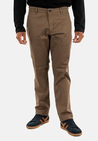 Man wearing brown straight-leg pants, black long-sleeve shirt, and black sneakers with brown stripes on a white background.