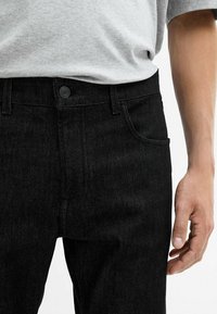Jeans in denim nero con vestibilità slim, dotati di tasca frontale, chiusura con bottoni e trama sottile. Indossati con una t-shirt grigia sopra.