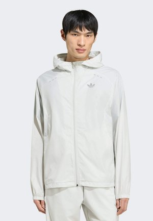 Jonge man draagt een witte lichtgewicht capuchonjas met een rits aan de voorkant en het Adidas-logo op de linkerborst.