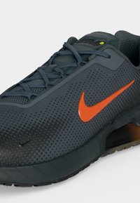 Σκούρο γκρι αθλητικό παπούτσι Nike με πορτοκαλί λογότυπο Swoosh, μαύρη σόλα και εμφανή αερόσολα, κεκλιμένο για να δείχνει το πλάι και τα κορδόνια.