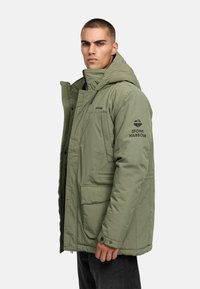 Khaki puffer jope kapuutsiga, millel on kaks esist pocketit, tõmblukuga sulgemine ja logod rinnal ja varrukal. Sujuv, isoleeritud kangas.