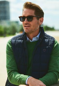 Marineblauwe puffer vest over een groene trui, witte geblokte shirt zichtbaar bij de kraag, bruine schildpadbril. Buitensetting met een vervaagd achtergrond.