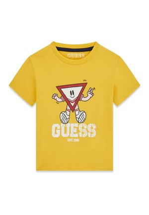 T-shirt jaune à manches courtes avec un personnage en forme de triangle rouge de dessin animé faisant un signe de paix, texte blanc « GUESS EST. 1981 » sous le personnage.