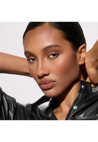 Gros plan sur un visage à la peau lisse, portant une veste en cuir noire brillante. Les traits comprennent des yeux marron, des lèvres pleines et des cheveux coiffés en arrière.