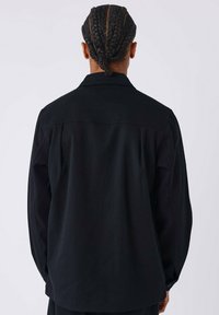Zwarte shirtjack met een relaxed fit, uitgerust met een puntige kraag, lange mouwen en een gladde textuur. De achterkant onthult subtiele stiksels.