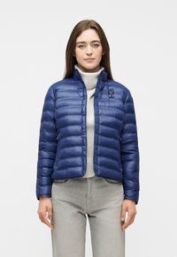 Γυναίκα που φοράει μπλε καπιτονέ μπουφάν πάνω από ανοιχτό γκρι πουλόβερ με ψηλό γιακά και ανοιχτό γκρι παντελόνι, στέκεται μπροστά σε απλό φόντο.