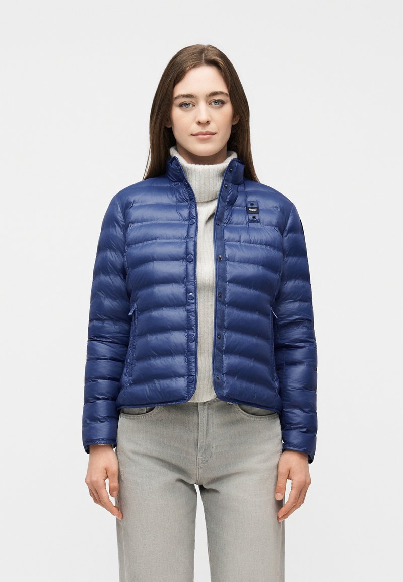 Γυναίκα που φοράει μπλε καπιτονέ μπουφάν πάνω από ανοιχτό γκρι πουλόβερ με ψηλό γιακά και ανοιχτό γκρι παντελόνι, στέκεται μπροστά σε απλό φόντο.