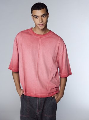 Camicia oversize a maniche corte in sfumatura di rosa, con scollo arrotondato e dettagli di cuciture visibili. Abbinata a pantaloni in denim scuro.