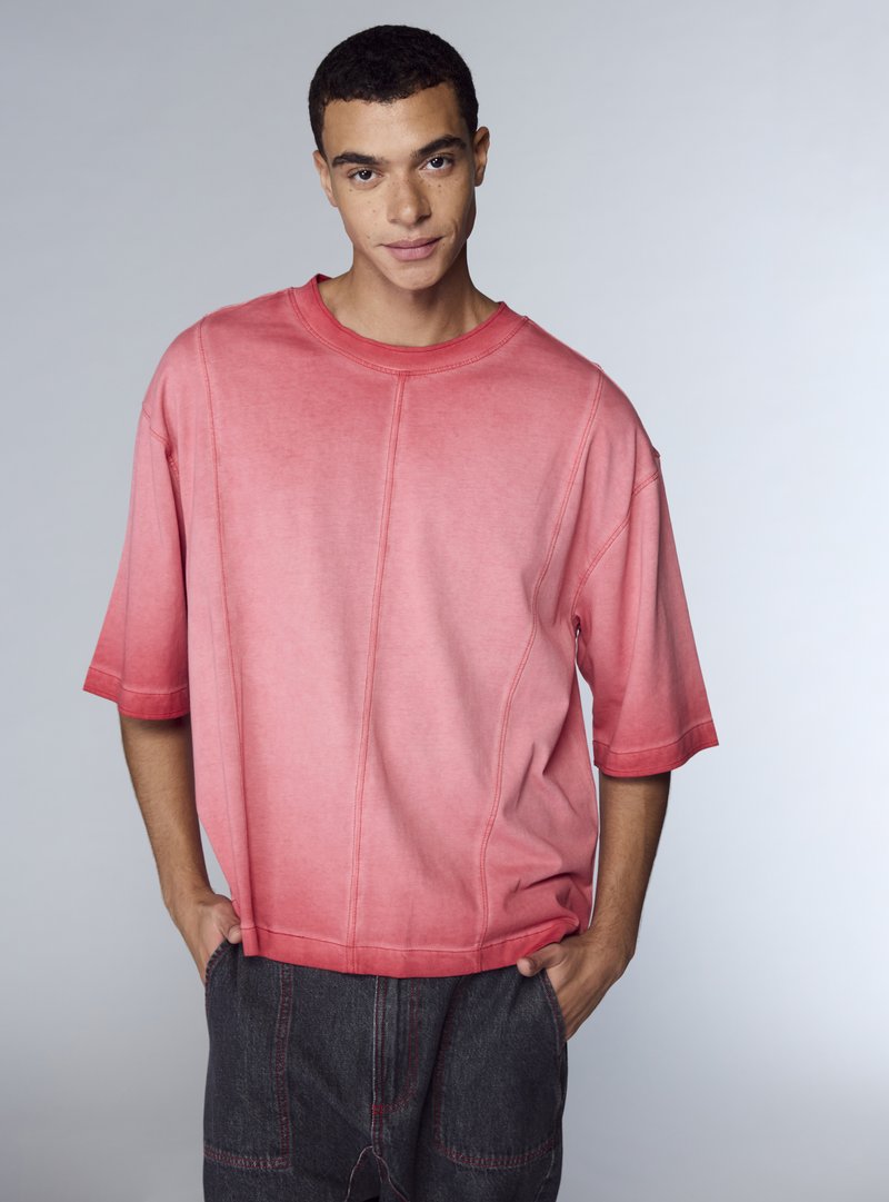 Camicia oversize a maniche corte in sfumatura di rosa, con scollo arrotondato e dettagli di cuciture visibili. Abbinata a pantaloni in denim scuro.