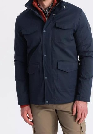 Mand iført navyjakke med klaplommer over en rød sweater og ternet skjorte, kombineret med beige cargobukser.