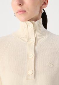 Pull en tricot couleur crème avec un col haut et cinq boutons visibles. Présente un logo subtil et une texture douce.