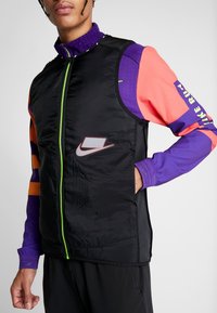 Homme portant une veste noire zippée avec une fermeture éclair vert néon et une chemise multicolore à manches longues avec des manches violettes, orange et rouges.