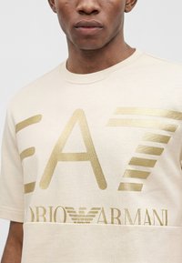 Άνδρας που φοράει μπεζ μπλουζάκι Emporio Armani EA7 με μεγάλο χρυσό λογότυπο στο στήθος, κομμένο από τον λαιμό και το μέσο του κορμού.