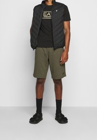 Svart vadderad väst över en svart t-shirt med guld logotyp, olivgröna shorts, svarta strumpor och svarta sandaler. En enkel och avslappnad look för aktiva kläder.