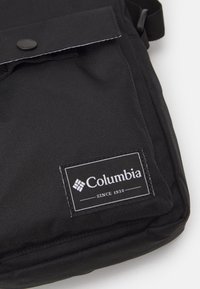 Borsa a messenger nera in nylon con chiusura a pattina, dotata di un pulsante circolare e di una toppa con logo bianco, che include il testo "SINCE 1938."