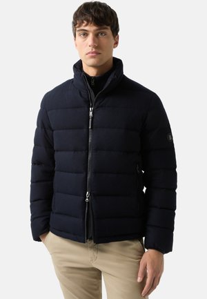 Opgepompte jas in donker marineblauw, met ritssluiting aan de voorkant, hoge kraag, doorgestikt ontwerp, met een logo-patch op de linkerarm, gedragen over een zwarte coltrui.