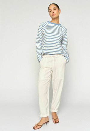 Žena oblečená v modro-bielom pruhovanom tričku s dlhým rukávom, krémových nohaviciach so sťahovacou šnúrkou a manžetami a hnedých sandáloch na podpätku, stojaca pred jednoliatym pozadím.