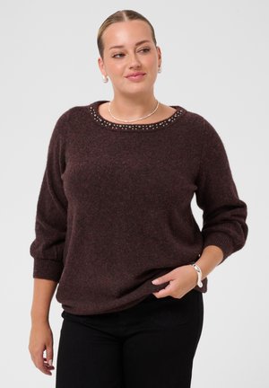 Femme portant un pull marron foncé avec un col clouté et un pantalon noir, tenant l'ourlet du pull d'une main, regardant légèrement sur le côté.
