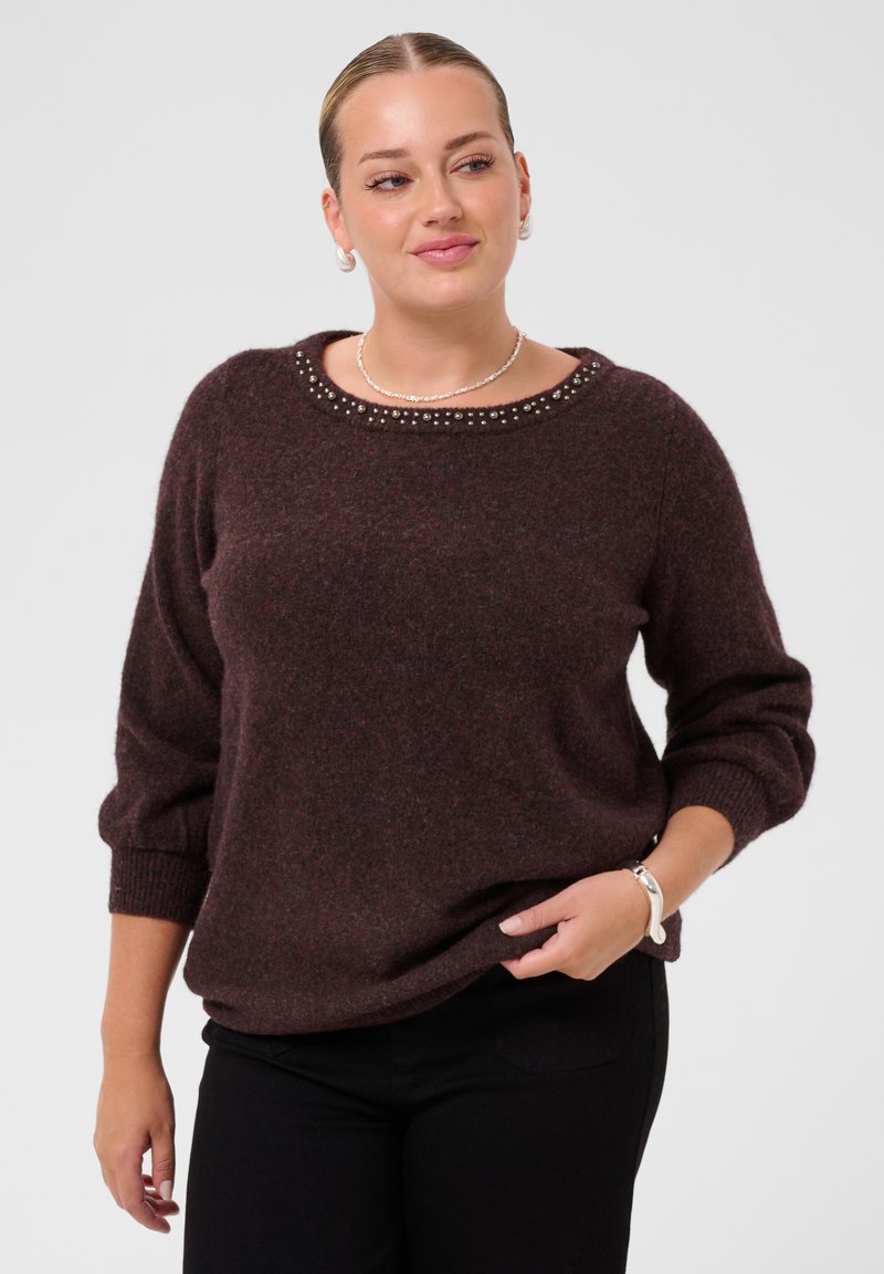 Femme portant un pull marron foncé avec un col clouté et un pantalon noir, tenant l'ourlet du pull d'une main, regardant légèrement sur le côté.