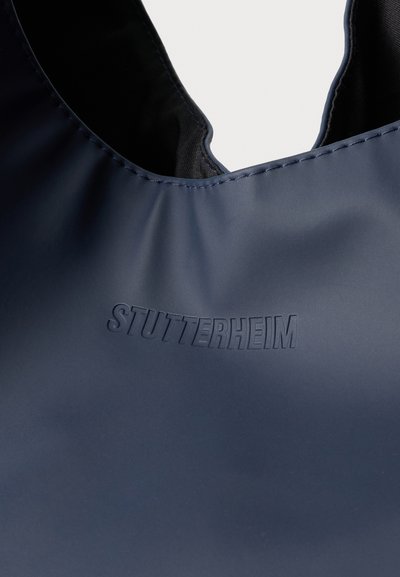 Granatowy płaszcz przeciwdeszczowy o gładkiej teksturze, z zakrzywionym dekoltem i wytłoczonym logo "STUTTERHEIM". Stębnowane wykończenia wzdłuż krawędzi.