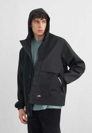 Junger Mann mit lockigem Haar, der eine schwarze Kapuzenjacke und ein grünes Hemd trägt, mit einer Hand in der Tasche stehend und zur Seite blickend.