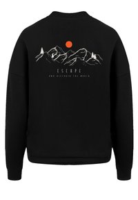 Sudadera negra con una escena de montaña y un sol rojo. El texto dice "ESCAPA Y DESCUBRE EL MUNDO" en blanco, sobre un fondo oscuro.